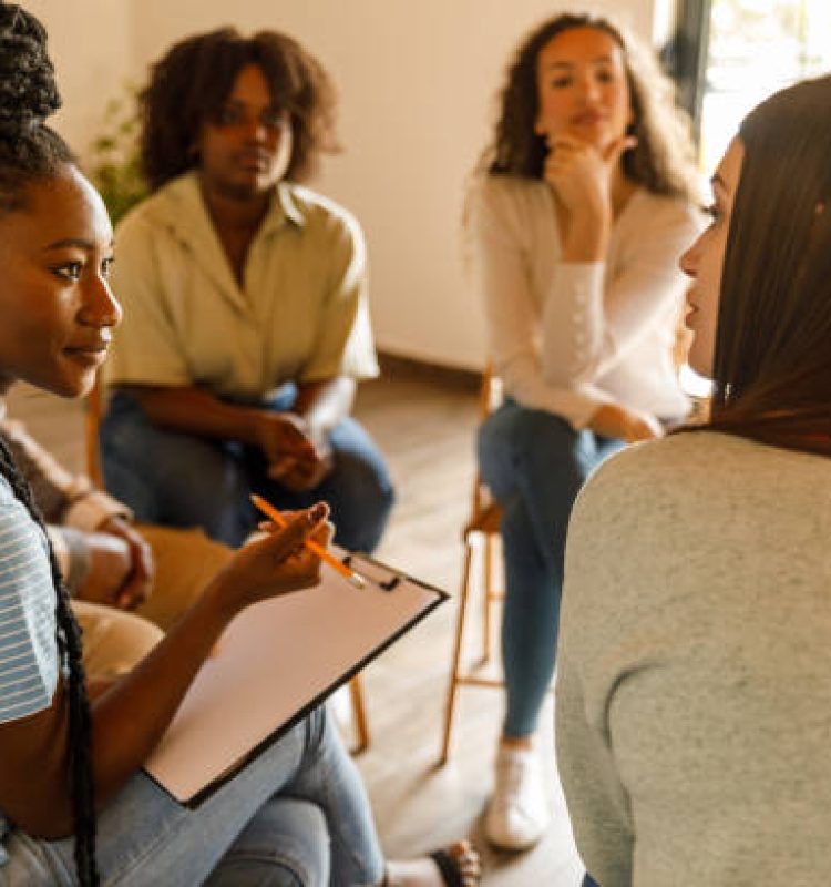 Selective focus shot of female therapist sitting in circle, holding a clipboard to take notes and talking during a group therapy session.